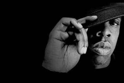 Black-and-white portrait of a man adjusting the brim of his cap against a dark background.