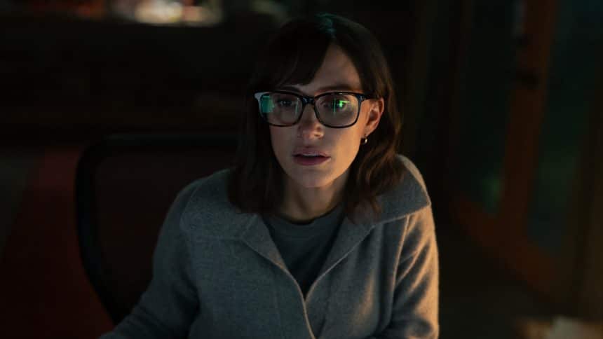 Woman with glasses focused on a computer screen in dim light.