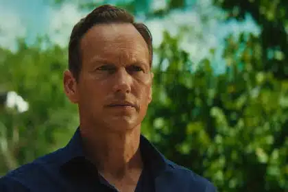 Man with a serious expression standing outdoors with trees in the background.