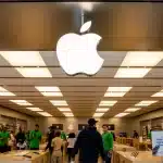 Interior of an Apple Store with customers and staff under a large Apple logo.