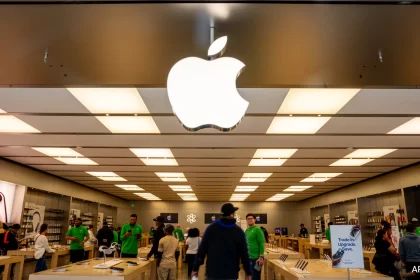 Interior of an Apple Store with customers and staff under a large Apple logo.