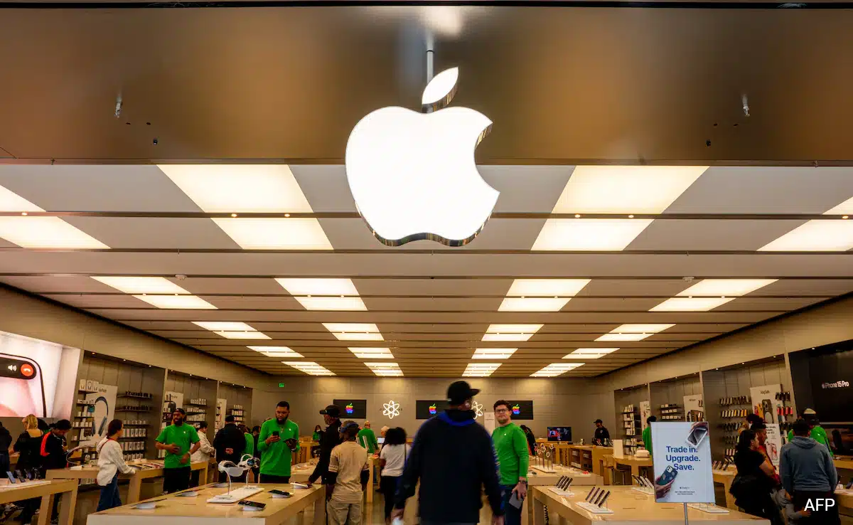 Interior of an Apple Store with customers and staff under a large Apple logo.
