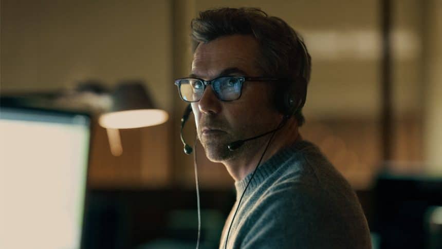 Patrick Brammall wearing a headset in a dimly lit office setting.