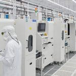 Workers in cleanroom suits operating machines in a high-tech manufacturing facility.